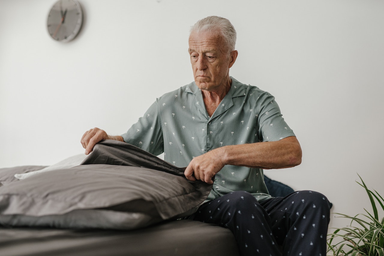 An Elderly Man in Gray Shirt and Black Pants Sitting on the Bed | Veteran Car Donations

