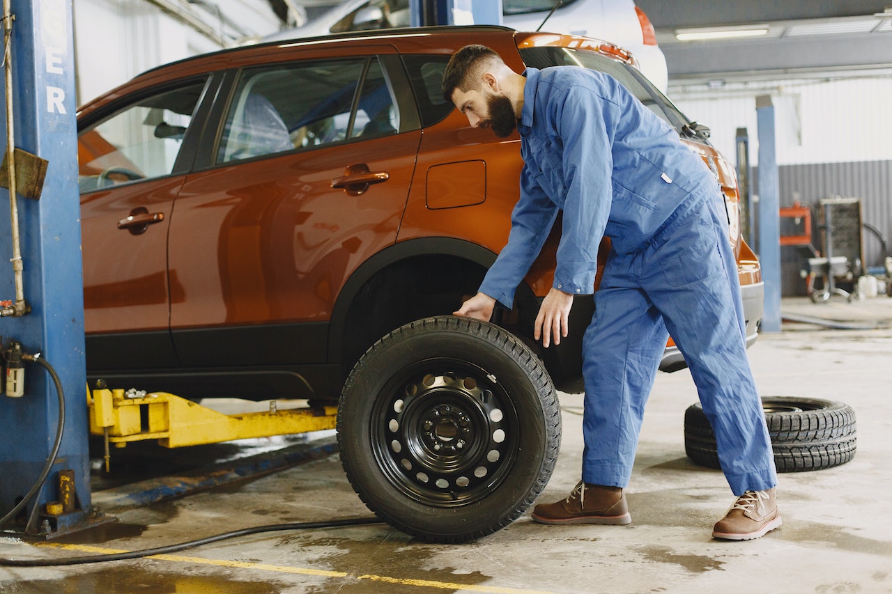 Man in Blue Coveralls Standing Beside an Orange Car Checking on the Tire | Veteran Car Donations
