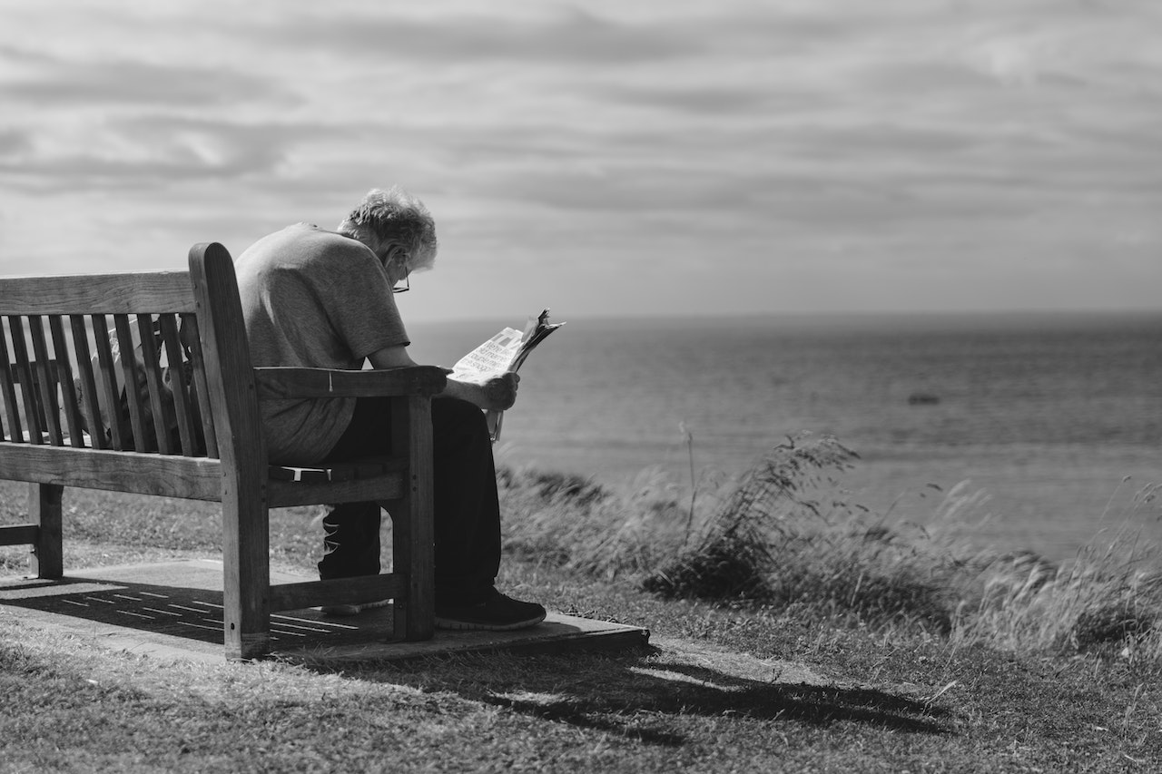 Grayscale Photo of Man Sitting on Brown Wooden Bench Reading News Paper during Day Time | Veteran Car Donations
