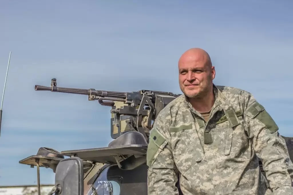 Man in US Army uniform sitting next to a machine gun mounted on a vehicle